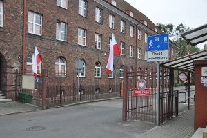 Główna brama wjazdowa do słupskiej Szkoły Policji. Na słupach trzymających skrzydła bramy zawieszone biało-czerwone flagi. Ujęcie z prawej strony.