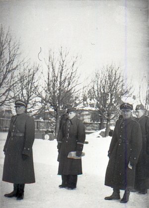 Grupa kilku oficerów stoi na placu w płaszczach. Jest zima. Jeden trzyma w ręku proporzec.